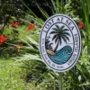 Hand-painted 'Lost at Sea' sign with a palm tree and ocean wave design, set in a natural garden with bright red flowers. The sign reads '528 East End Rd., St. Croix, Virgin Islands' around the border.