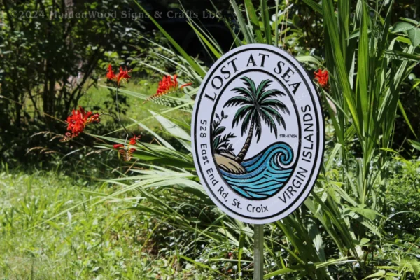 Hand-painted 'Lost at Sea' sign with a palm tree and ocean wave design, set in a natural garden with bright red flowers. The sign reads '528 East End Rd., St. Croix, Virgin Islands' around the border.